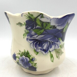 May include: A white ceramic planter with a scalloped rim features a blue and green floral pattern of roses.