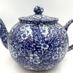 May include: A round ceramic teapot featuring a deep blue base colour with a repeating white floral and vine pattern. The teapot includes a matching lid with a small knob handle, a curved spout, and a sturdy side handle, all covered in the same floral design.
