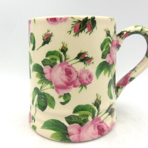 Könnte beinhalten: Eine weiße Keramiktasse mit einem rosa-grünen Blumenmuster. Die Tasse hat einen Henkel und ist mit rosa Rosen und grünen Blättern verziert.