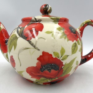 May include: A ceramic teapot with a poppy floral design. The teapot has a cream-coloured base decorated with red poppies, green leaves, and black accents. The handle, spout, and lid also feature the poppy pattern.