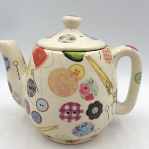 May include: A white ceramic teapot with a colourful pattern of buttons, thread, scissors, and other sewing-themed imagery. The teapot has a lid and a handle.