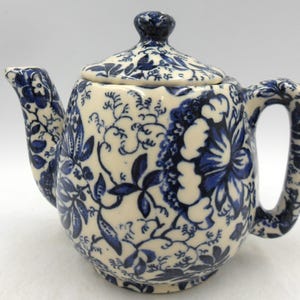 Könnte beinhalten: Eine weiße Teekanne mit einem blauen Blumenmuster. Die Teekanne hat einen Deckel und einen Henkel.