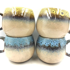 May include: Four ceramic mugs stacked in two pairs. The top pair has a yellow, brown, and tan glaze, while the bottom pair is blue, brown, and tan. Each mug has a dark brown handle.