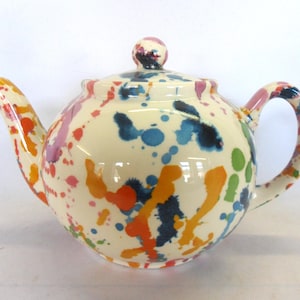 May include: A white ceramic teapot with a colourful splatter paint design. The teapot has a round body, a spout, a handle, and a lid. The colours in the design include orange, yellow, blue, green, and pink.