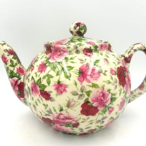 Peut inclure: Une théière en céramique blanche avec un motif floral de roses roses et rouges. La théière a un corps rond et un bec verseur sur le côté gauche.