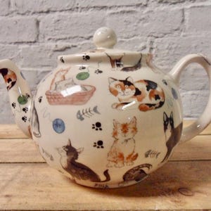 May include: A white ceramic teapot featuring a cat design. The teapot has a rounded body and a curved spout. The handle is on the right side of the teapot. The teapot is decorated with a pattern of cats, balls of yarn, and fish bones.