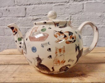 May include: A white ceramic teapot featuring a cat design. The teapot has a rounded body and a curved spout. The handle is on the right side of the teapot. The teapot is decorated with a pattern of cats, balls of yarn, and fish bones.