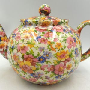 May include: A floral patterned ceramic teapot. The teapot features a rounded body, a curved spout, and a loop handle. The lid is topped with a flower-shaped knob. The pattern includes pink, yellow, orange, blue, and white flowers.