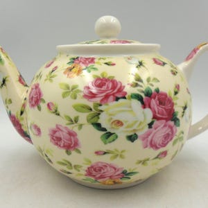 May include: A white ceramic teapot with a floral pattern of pink and white roses on a cream background. The teapot has a lid with a knob on top.