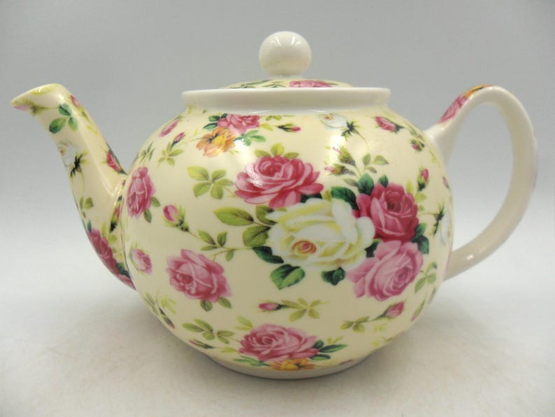 May include: A white ceramic teapot with a floral pattern of pink and white roses on a cream background. The teapot has a lid with a knob on top.