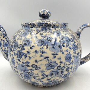 May include: A round ceramic teapot with a lid, spout, and handle. The teapot is decorated with a blue floral pattern on a cream background. The lid has a small knob. The teapot is designed for brewing tea.