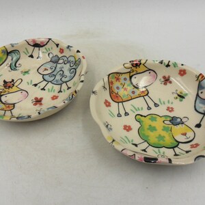 May include: Two small ceramic dishes with a white background and colourful cartoon sheep and cow designs. The dishes are ideal for serving snacks or condiments.