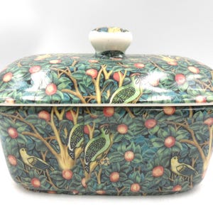 May include: A rectangular ceramic butter dish with a lid featuring a detailed pattern of green trees, orange fruit, and birds. The lid has a white knob handle on top. The overall design displays a dense botanical motif with birds perched.