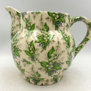 May include: Cream-colored ceramic pitcher with a rounded body and a curved handle. The pitcher is decorated with a pattern of green leaves, white flowers, and small purple flowers. The pitcher has a spout for pouring.