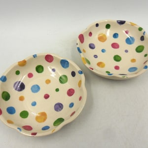 May include: Two small, round ceramic bowls with scalloped edges. The bowls have an off-white base colour and are decorated with hand-painted, multi-coloured polka dots in shades of blue, green, yellow, purple, and pink scattered across the surface.