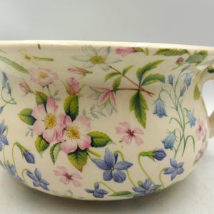以下が含まれることがあります： クリーム色のセラミック製おまる。ピンク、青、白の花と緑の葉が描かれています。側面には取っ手があり、縁が広がっています。