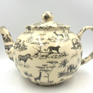 May include: A cream-coloured teapot with a black and white pattern of African animals, including zebras, giraffes, elephants, and lions. The teapot has a lid and a handle.