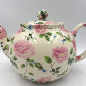 May include: A cream-coloured ceramic teapot decorated with pink roses, green leaves, and small blue birds. The teapot has a rounded body, a curved handle, and a lid with a decorative knob.