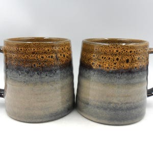 May include: A trio of ceramic mugs with a speckled, matte finish in cream, light grey, and dark grey. Each mug features a simple, modern design and a rounded handle.