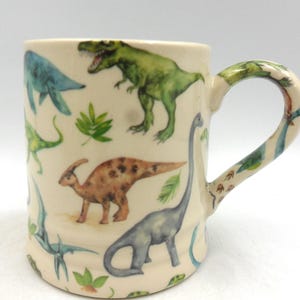 May include: A white ceramic mug with a dinosaur pattern. The mug features a variety of dinosaurs in different colours, including green, blue, brown, and orange. The dinosaurs are depicted in a whimsical style, with some of them having playful expressions.