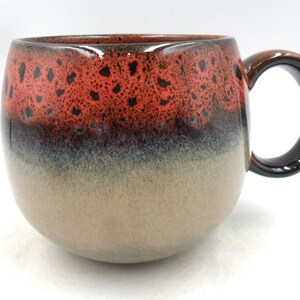 May include: A ceramic mug with a rounded shape and a large handle. The top of the mug is a speckled red, transitioning to a dark brown band, then a beige base. The handle is dark brown and black.