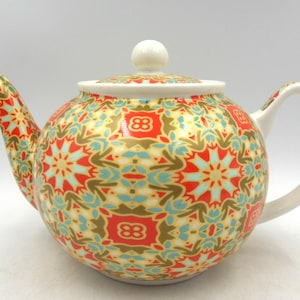 May include: A white ceramic teapot with a geometric pattern in shades of red, blue, and gold. The teapot has a round body and a white lid.