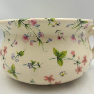 Peut inclure: Pot de chambre en céramique crème avec une anse, orné d'un motif floral de fleurs roses, violettes et bleues, ainsi que de feuilles vertes et de petits papillons. Le pot a une forme arrondie et un bord légèrement évasé.