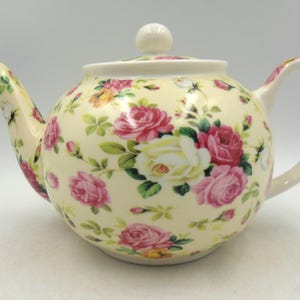 Könnte beinhalten: Eine cremefarbene Teekanne mit einem Blumenmuster aus rosa und weißen Rosen. Die Teekanne hat einen Deckel mit einem Knopf oben.