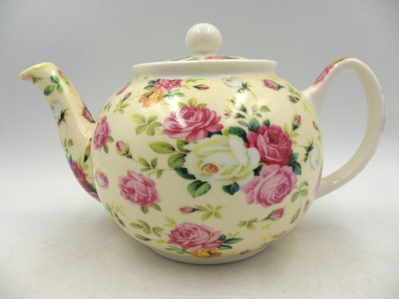 May include: A cream-colored teapot with a floral pattern of pink and white roses. The teapot has a lid with a knob on top.
