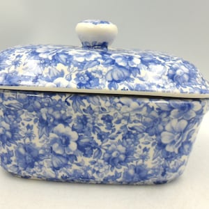 以下が含まれることがあります： 青と白の花柄が施された長方形の陶器製バターケース。上部には小さな丸い取っ手が付いた蓋が付属しています。全体に高密度で繰り返される植物のプリントデザインが施されており、表面は滑らかな質感で仕上げられています。