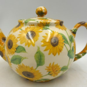 May include: A cream-coloured ceramic teapot adorned with yellow sunflowers and green leaves. The teapot features a rounded body, a curved spout, and a handle. The lid is topped with a small sunflower-shaped knob. The design is repeated on the spout and handle.