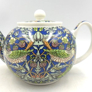 May include: A ceramic teapot featuring a blue background with a detailed pattern of birds, strawberries, and foliage. The teapot has a white handle, spout, and lid with a small white knob. The design is symmetrical and ornate.