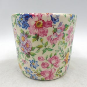 May include: A ceramic cup with a floral design. The cup has a cream-coloured base decorated with pink, blue, and yellow flowers, and green leaves. The flowers are in full bloom, creating a vibrant and detailed design.