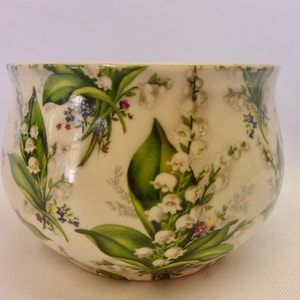 May include: A white ceramic bowl decorated with a floral pattern of lily of the valley, green leaves, and small blue and red flowers. The bowl has a rounded shape and a slightly flared rim.
