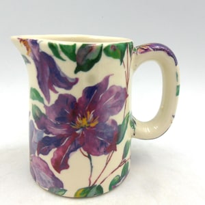 Könnte beinhalten: Eine weiße Keramikkanne mit Blumenmuster. Die Kanne hat ein violettes und grünes Blumenmuster auf weißem Hintergrund. Die Kanne hat einen Henkel und einen Ausguss.