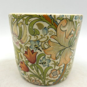 Peut inclure: Une tasse en céramique avec un motif floral dans des tons de vert, orange, jaune et bleu. Le motif représente des lys stylisés et d'autres fleurs.