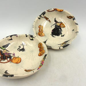 May include: Two white ceramic Halloween-themed bowls with black and orange designs. One bowl features a witch riding a broomstick and the other bowl features a black cat and pumpkins.