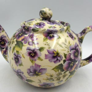 以下が含まれることがあります： クリーム色の陶器のティーポット。紫色のパンジーと緑の葉のフローラルパターンで飾られています。 ティーポットは丸みを帯びたボディ、湾曲したハンドル、小さなノブが付いたお揃いの蓋が特徴です。
