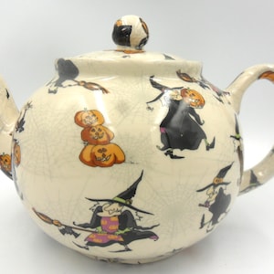 May include: A white ceramic teapot with a black and orange Halloween design. The teapot features witches, cats, and pumpkins.