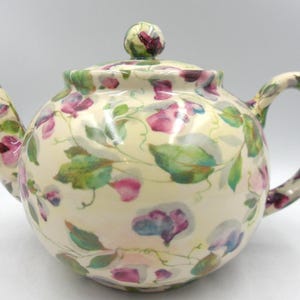 May include: A cream-coloured ceramic teapot with a floral design. The teapot has a rounded body, a curved handle, and a spout. The floral pattern includes pink, purple, and green flowers and leaves. The lid has a small, round knob.