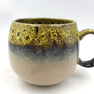 Peut inclure: Une tasse en céramique de forme arrondie avec une grande anse. Le haut de la tasse est jaune avec des mouchetures noires, passant au gris puis à une base beige. L'anse est marron foncé.
