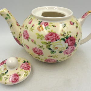 May include: A white ceramic teapot with a floral pattern in shades of pink, yellow, and green. The teapot has a lid and a handle. The teapot is sitting on a white surface.
