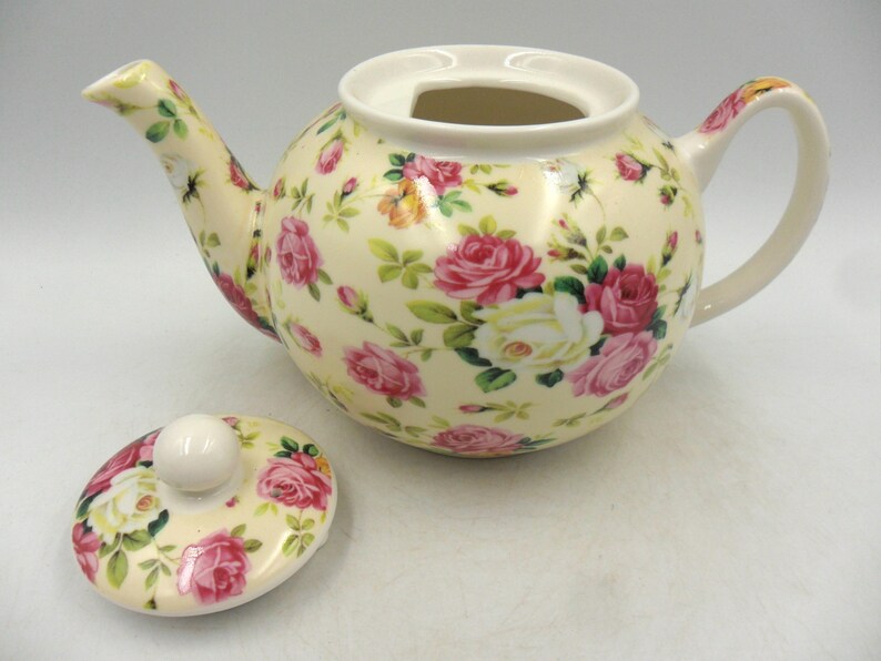 May include: A white ceramic teapot with a floral pattern in shades of pink, yellow, and green. The teapot has a lid and a handle. The teapot is sitting on a white surface.