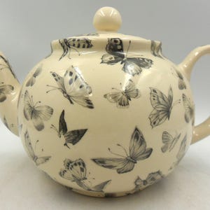 May include: Cream-coloured ceramic teapot with a lid and handle. The teapot is decorated with a pattern of black and grey butterflies. The spout and handle are curved, and the lid has a small knob.