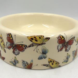 May include: A white ceramic bowl with a butterfly design. The bowl features colourful butterflies with red, yellow, blue, and brown accents.