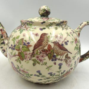 May include: Cream-coloured ceramic teapot with a lid and handle. The teapot is decorated with images of birds, flowers, and berries. The spout and handle have detailed floral patterns. The lid has a small, round knob.