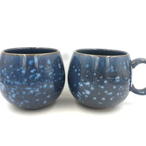 May include: Two blue ceramic mugs with a speckled glaze. The mugs have a rounded shape and a handle.