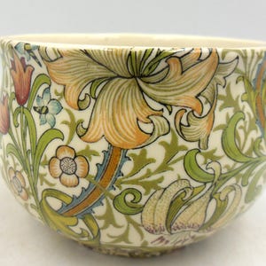 May include: A ceramic bowl with a floral design. It features large orange and yellow lilies, green leaves, and smaller flowers in red, blue, and orange against a cream background. The bowl has a slightly flared rim.