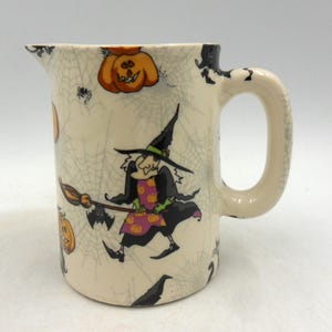 May include: A white ceramic jug with a black handle. The jug is decorated with a Halloween theme, featuring witches, pumpkins, and cobwebs.