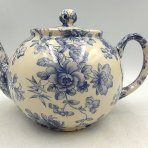 May include: A white teapot with a blue floral pattern. The teapot has a round body and a curved spout. The handle is also curved and has a floral design.